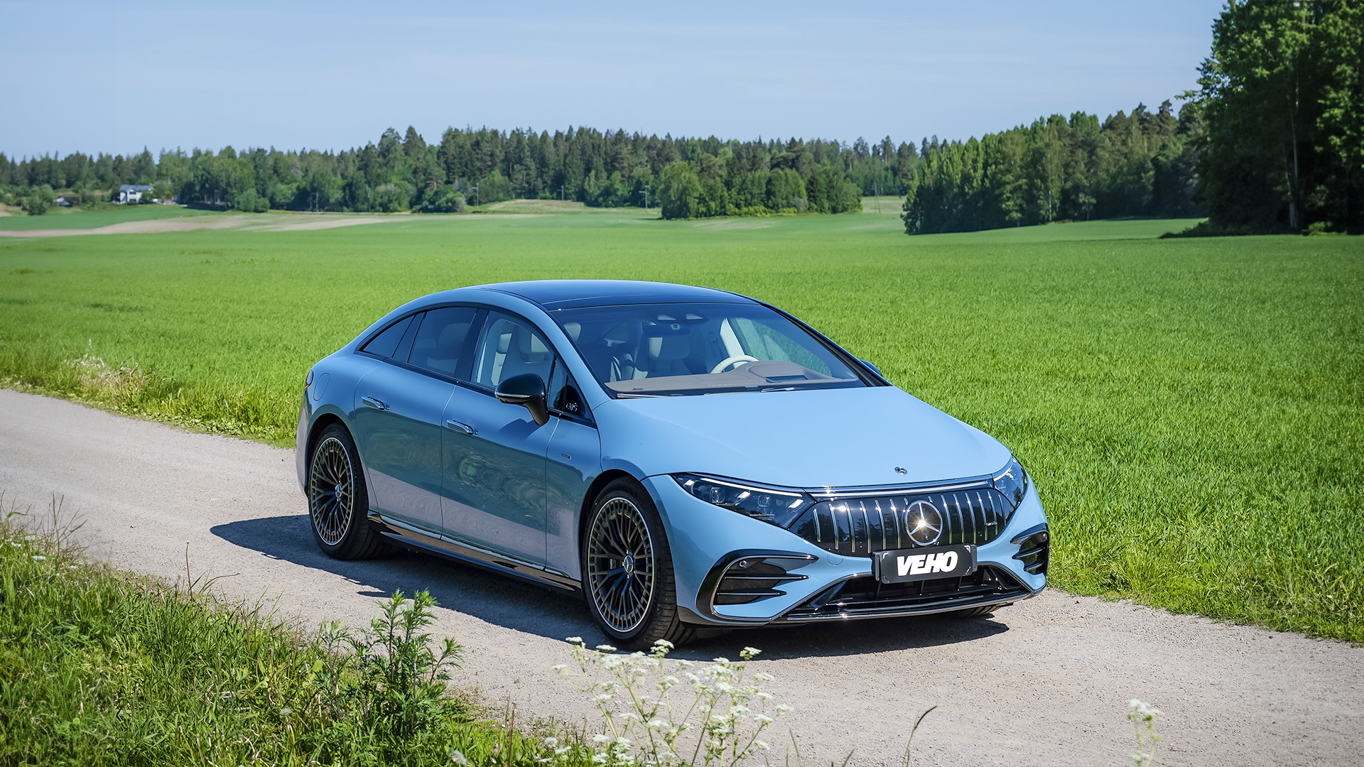 Vaaleansininen henkilöauto hiekkatiellä vihreän pellon laidalla.