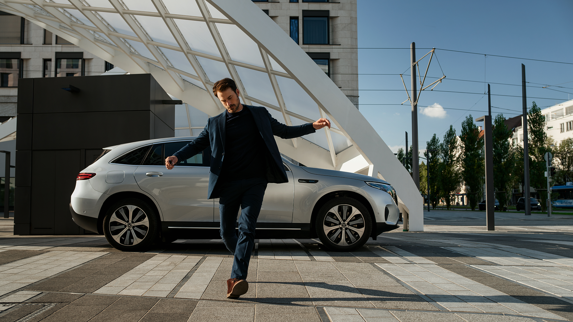 Mies tanssahtelee vapautuneesti kadulla kaupunkinäkymässä. Taustalla hopeinen Mercedeksen maasturi.