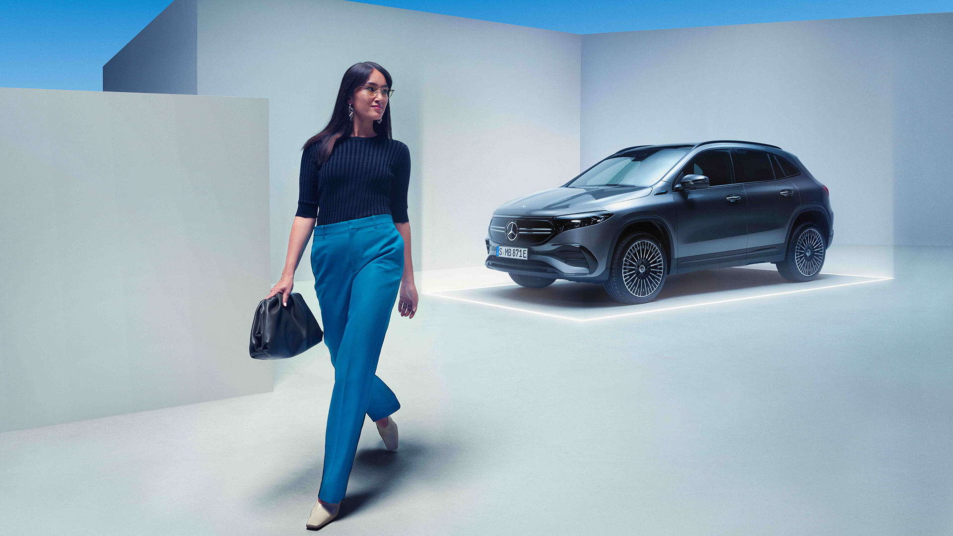 Henkilö kävelee kohti kameraa pelkistetyn valkoist ataustaa vasten. Taustalla uusi Mercedes-täyssähköauto.