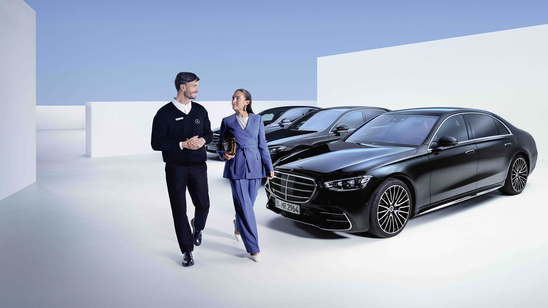 Kaksi henkilöä kävelee pelkistetyssä valkoisessa ympäristössä, jossa useita mustia Mercedeksiä taustalla.