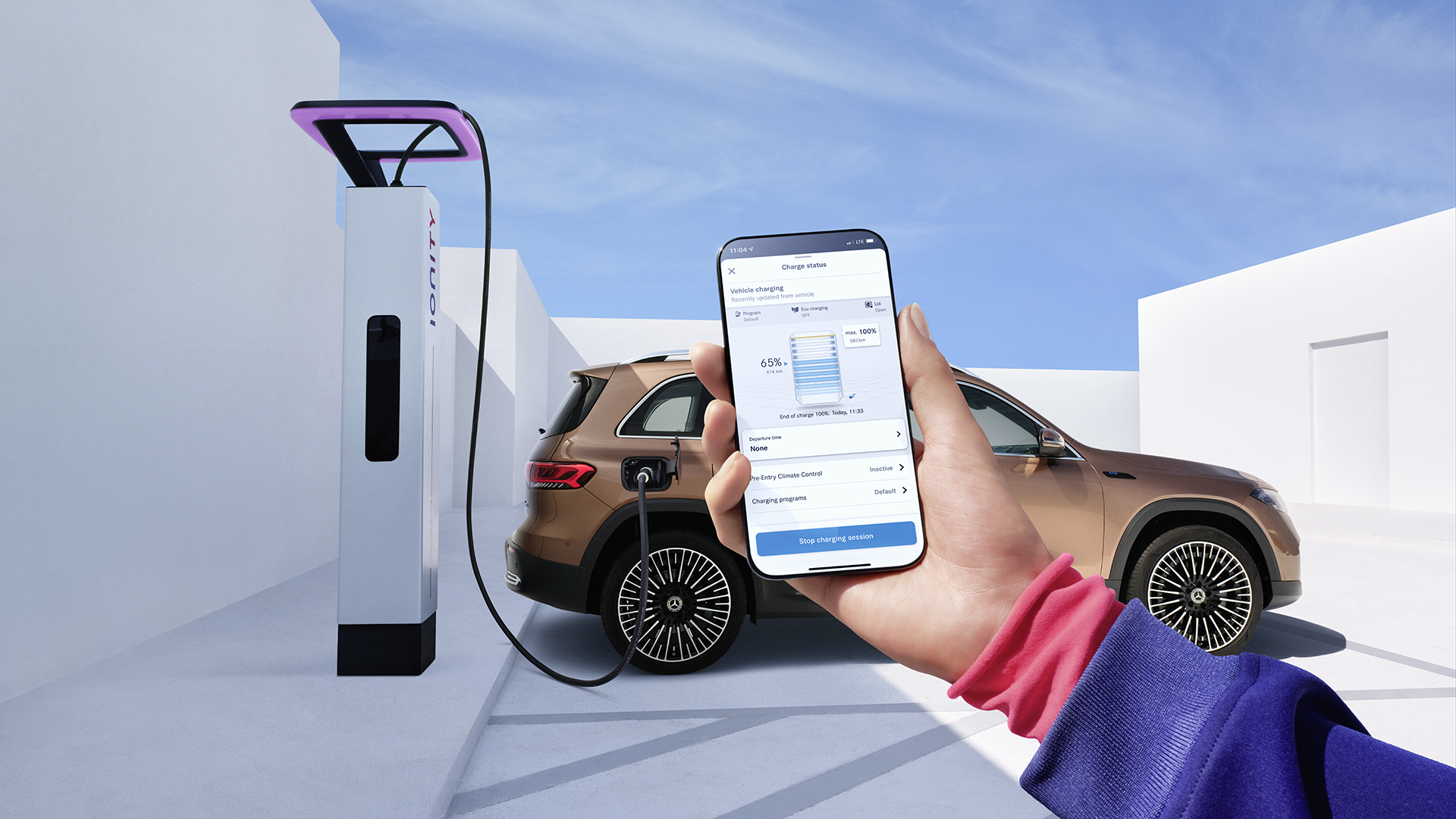 Käsi, jossa puhelin. Puhelimessa avoinna Mercedes me Charge-sovelluksen latausnäkymä. Taustalla kuparin värinen Mercedes EQB latauksessa.