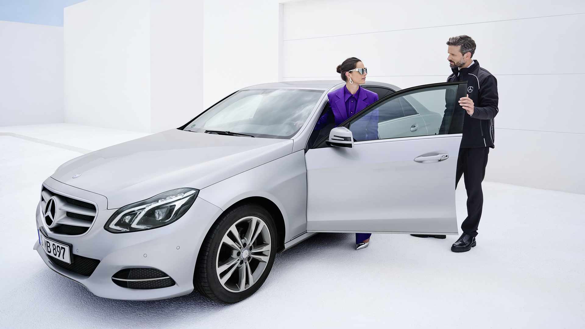 Asiakas ja asiakaspalvelija hopeanharmaan Mercedes-Benz E-sarjan vieressä.